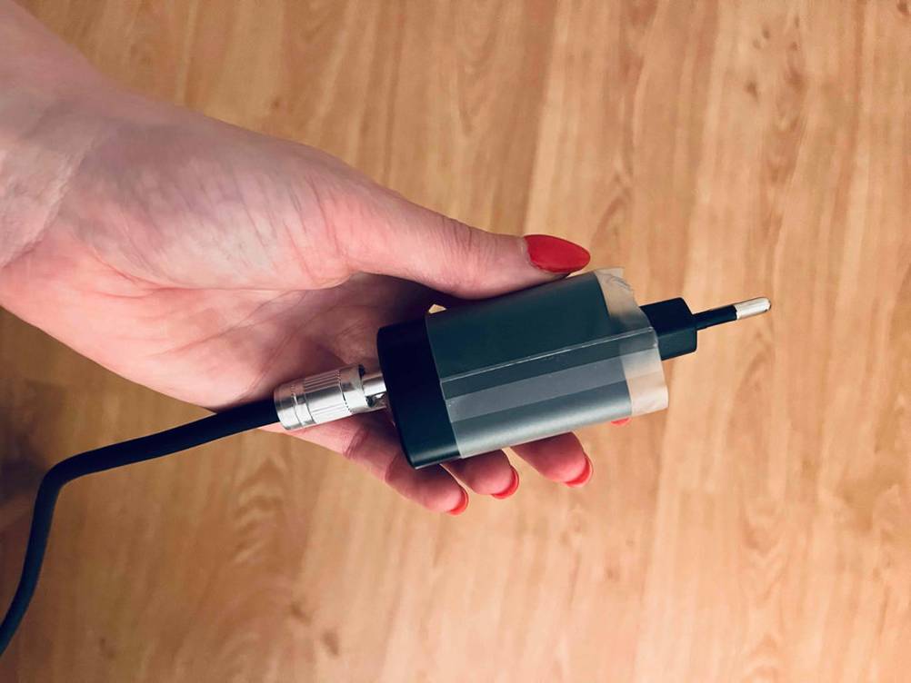 Lötkolben im Test: Nahaufnahme einer Hand mit Stecker-Adapter vor einem Holzhintergrund.