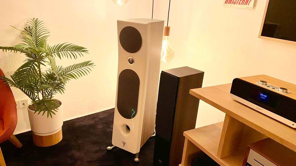 Hifi-Lautsprecher-Test: ein weißer und ein Standlautsprecher aus Holz stehen in einem Wohnbereich zwischen einer Topf-Palme und einem Holztisch mit Hifi-Geräten.