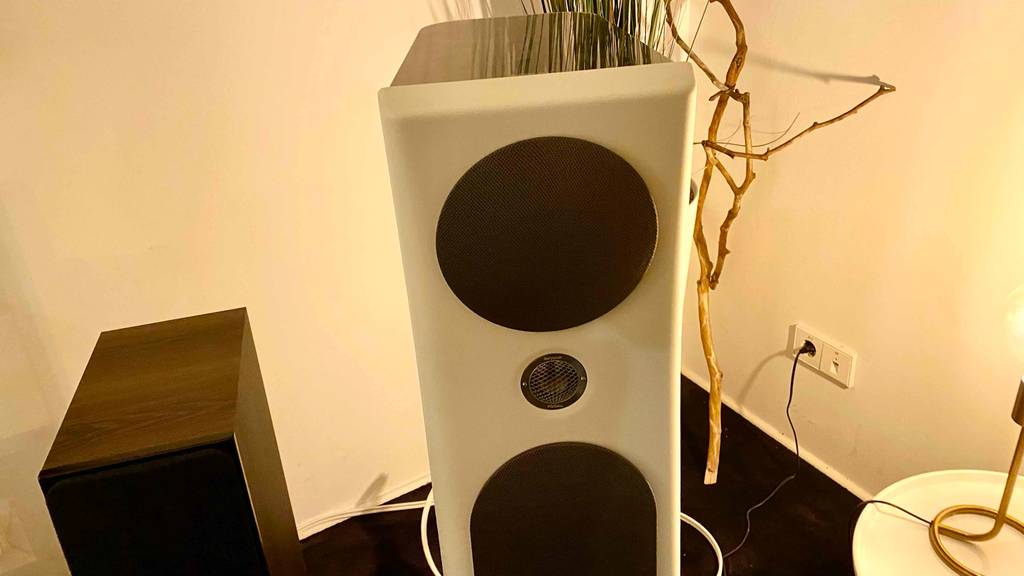 Hifi-Lautsprecher im Test: ein weißer und ein Standlautsprecher aus Holz stehen in einem Wohnbereich auf einem schwarzen Teppichboden.