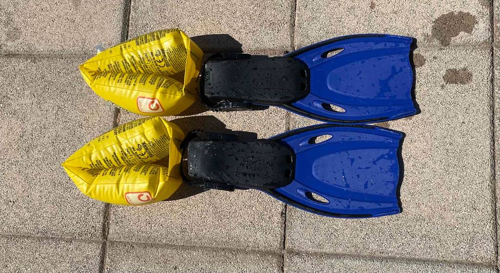 Schwarz-dunkelblaue Kinder-Schwimmflossen im Test liegen neben gelben Schwimmärmeln auf Zementpflastersteinen.