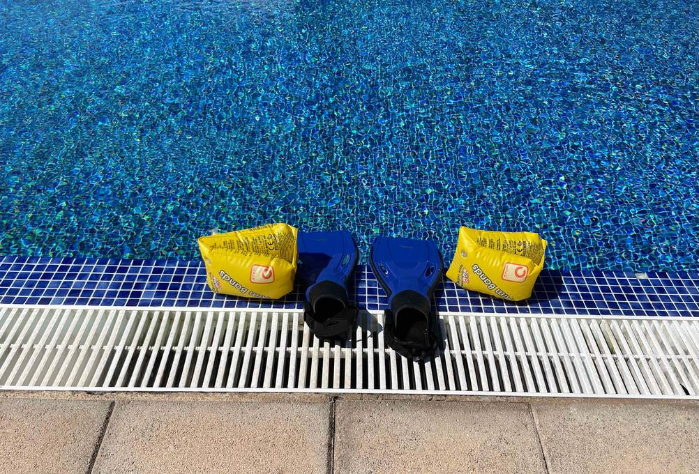 Schwimmflossen für Kinder im Test: blaue Schwimmflossen mit schwarzem, geschlossenen Zehenbereich stehen zwischen gelben Schwimmflügeln am Beckenrand eines Swimmingpools.