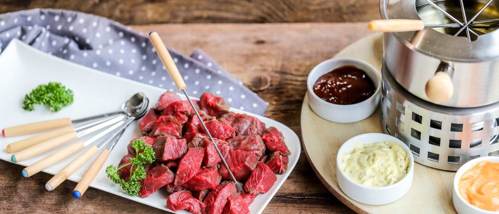 Fondue-Test: Ein Teller mit kleinen Fleischstücken, Fondue-Besteck und einem Fondue-Set stehen auf einem Tisch. 
