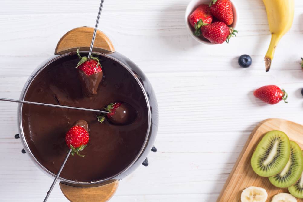Fonduegabeln-Test: In ein Schokoladenfondue werden drei Edelstahlgabeln mit einer Erdbeere eingetaucht. Daneben steht ein Brett mit Kiwis und Bananen und eine kleine Schale mit Erdbeeren. 