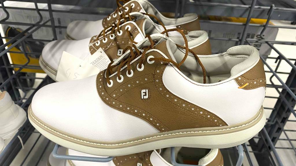 Golfschuhe im Test: Nahaufnahme zweifarbiger braun-weißer Golfschuhe aus Leder mit Schnürung, die sich aufgereiht in einem Ausstellungsregal befinden.