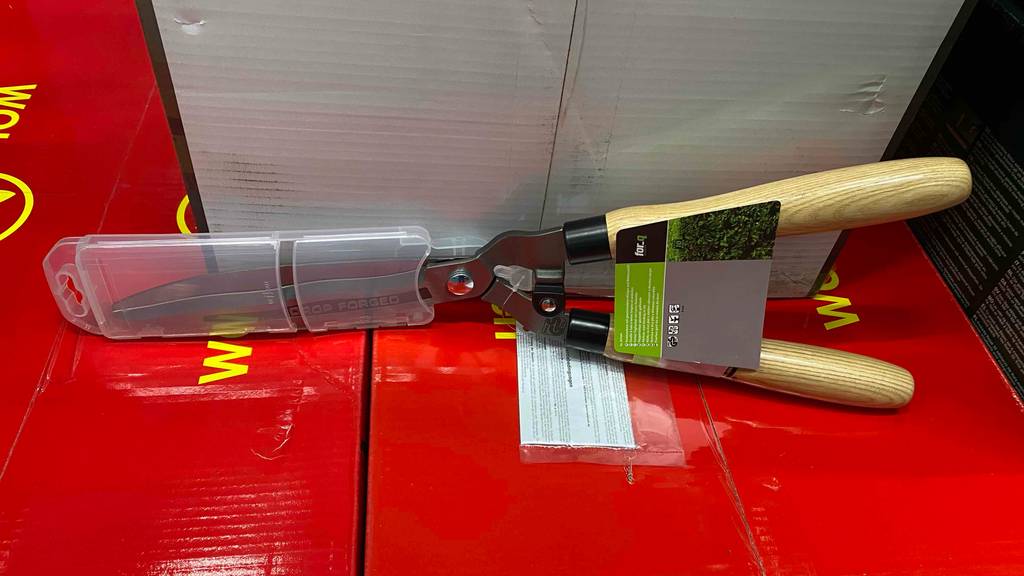 Im Test: Eine Hand-Heckenschere mit Holzgriff auf einem roten Karton lehnt an einer grauen Verpackung.