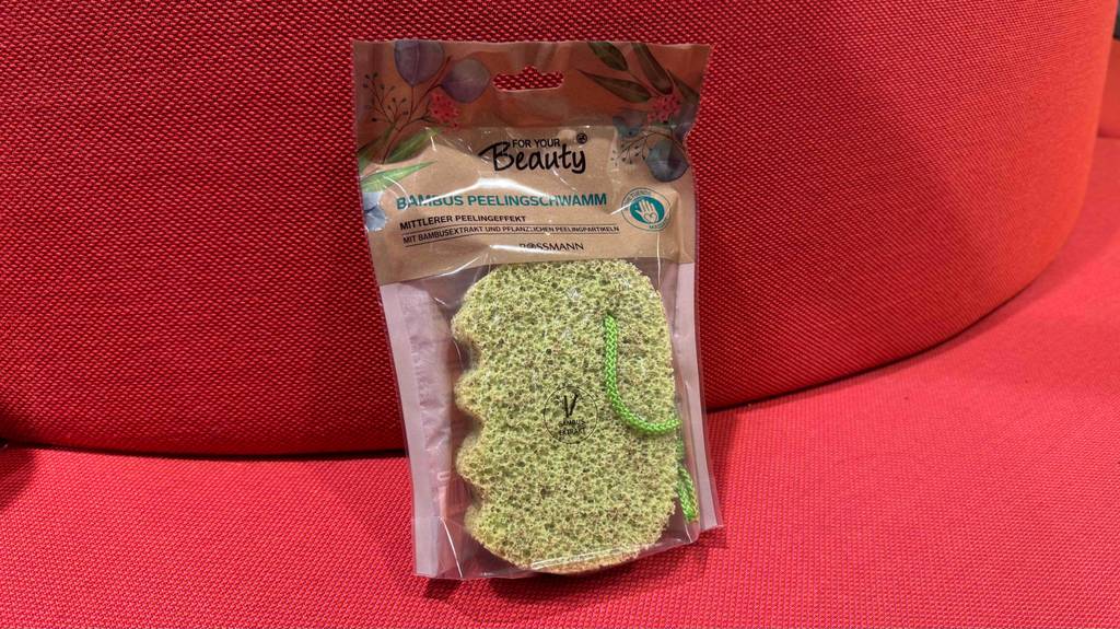 Ein in Plastik verpackter Peelingschwamm lehnt zum testen an ein rotes Polster.