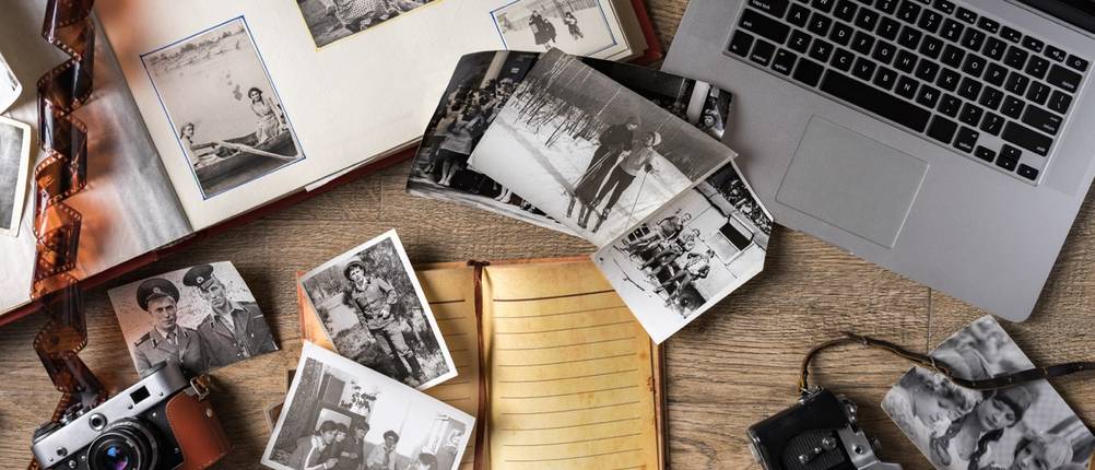 Mehrere ältere Fotoabzüge liegen neben Kamera und Computer.