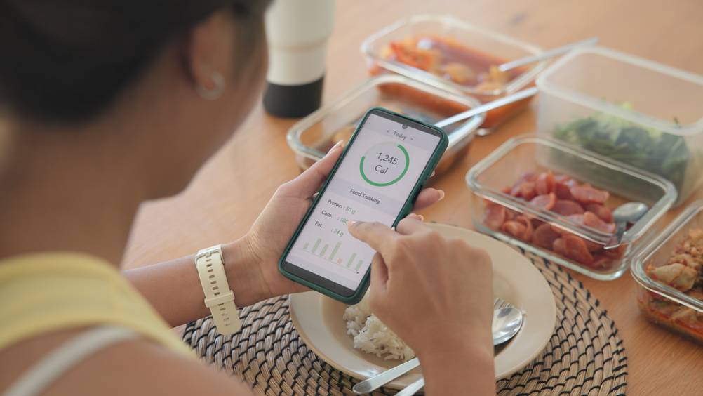 Fitnesstracker mit Blutdruckmessung im Test: Eine Frau checkt ihren Kalorienverbrauch auf dem Handy. Vor ihr auf dem Tisch stehen Glasbehälter mit Gemüse.