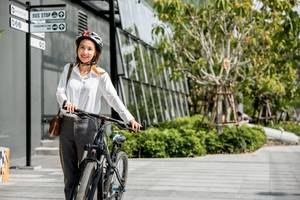 Eine Frau trägt ein Business-Outfit, einen Helm und schiebt ein Fahrrad auf dem Gehweg entlang.