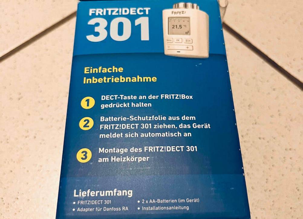 Smart-Home-Heizkörperthermostate im Test: eine blaue Verpackung mit gedruckter Anleitung zur Inbetriebnahme eines weißen Fritz!Dect 301 liegt auf beigefarbenen Fliesen.