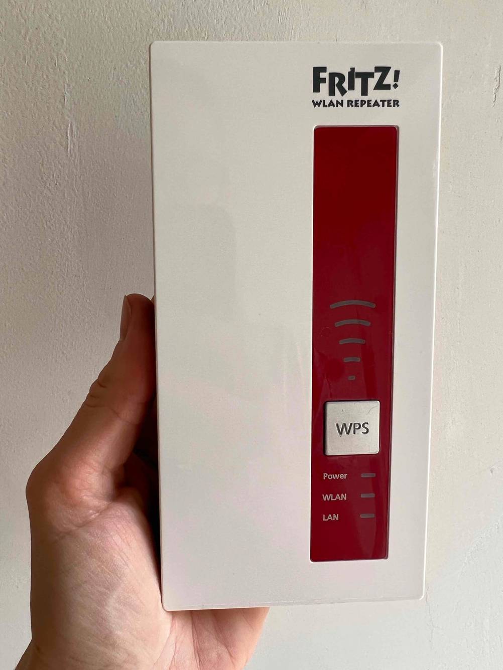 Hand hält einen getesteten WLAN Repeater von Fritz vor weißer Wand.