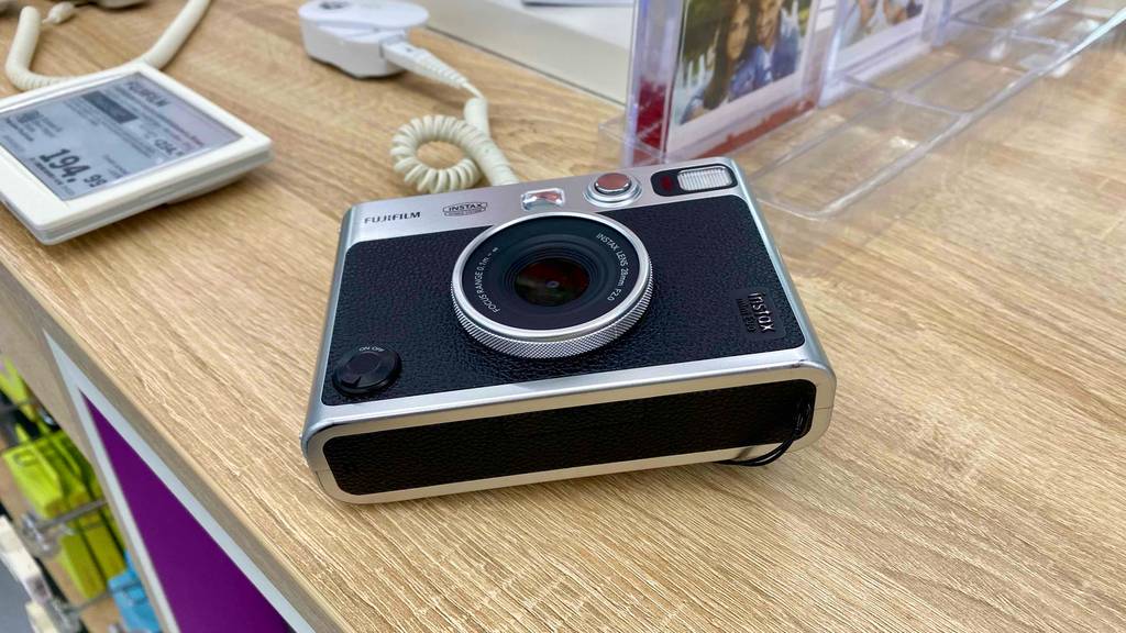 Ansicht von oben auf eine schwarz-silberne Fujifilm Sofortbildkamera im Test, die inklusive Preisschild gesichert auf einem Ausstellungstisch liegt.