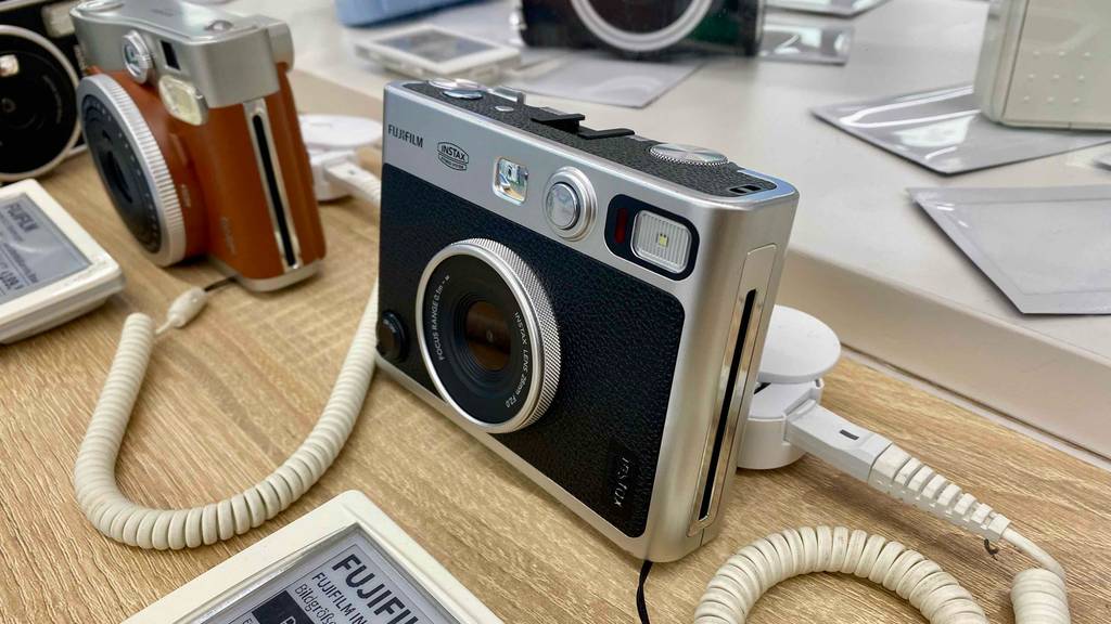 Seitliche Vorderansicht ein schwarz-silbernen Fujifilm Sofortbildkamera im Test, die gesichert auf einem Verkaufstisch ausgestellt ist.