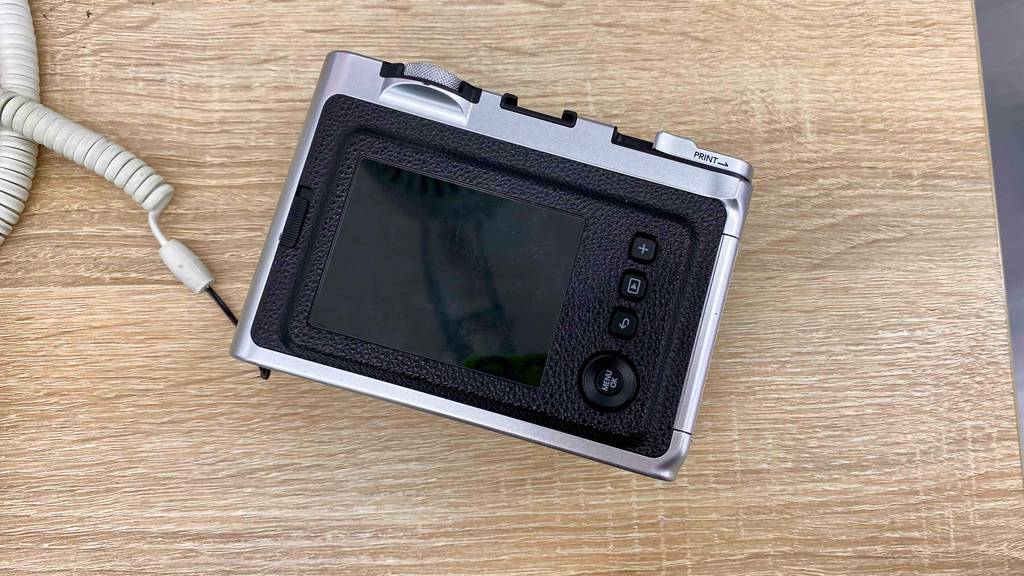 Ansicht des rückwärtigen Displays einer schwarz-silbernen Fujifilm Sofortbildkamera im Test, die gesichert auf einem Holztisch liegt.