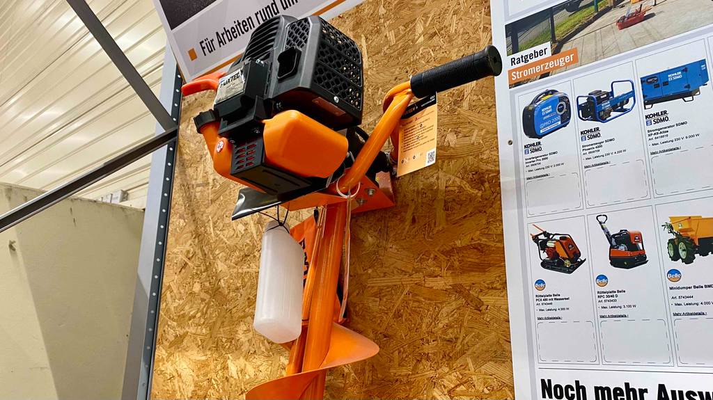Erdbohrer-Test: ein orangefarbener Erdbohrer mit Griff der Marke Fuxtec hängt an einem Ausstellungsstand, mit Fokus auf die Motoreinheit.
