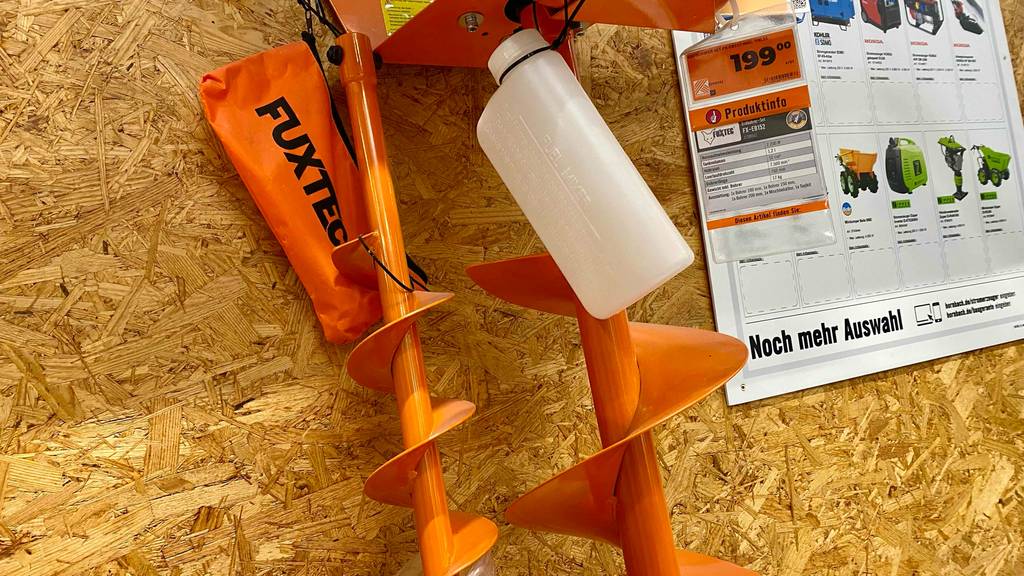 Erdbohrer-Test: ein orangefarbener Erdbohrer der Marke Fuxtec inklusive Zubehör hängt an einem Ausstellungsstand.