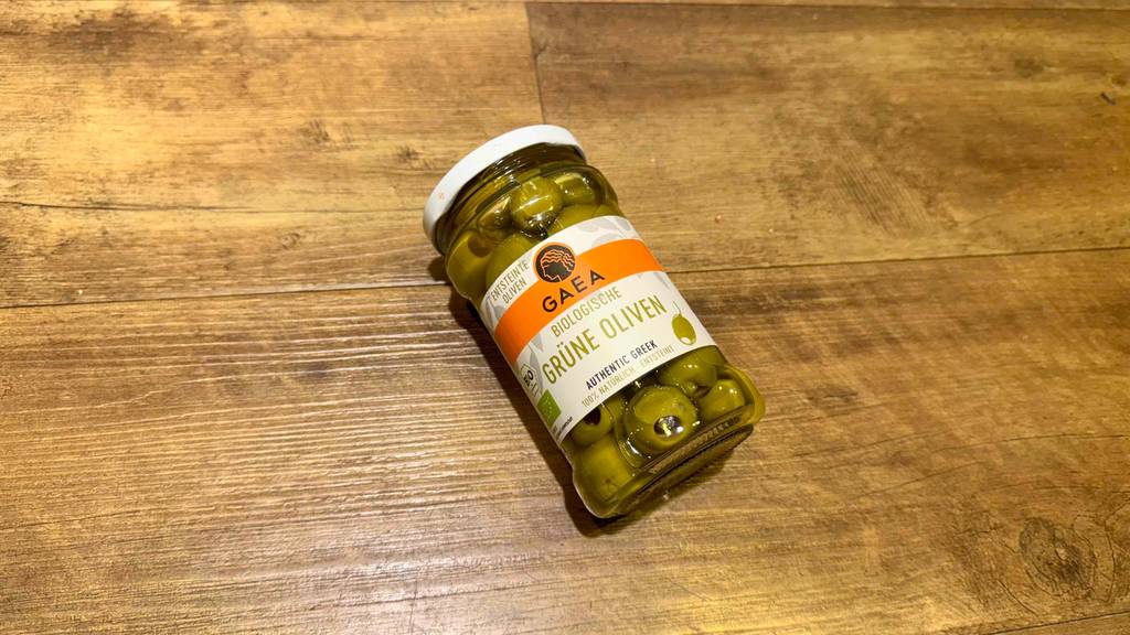 Im Test: Grüne Oliven im Glas liegen auf einer Holzoberfläche.