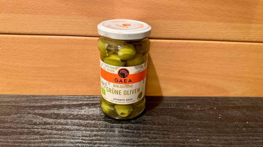 Getestet: Grüne Oliven im Glas stehen vor einer Holzfläche.