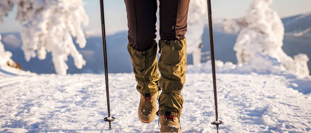 Eine Person mit Gamaschen und Wanderstöcken wandert im Schnee.