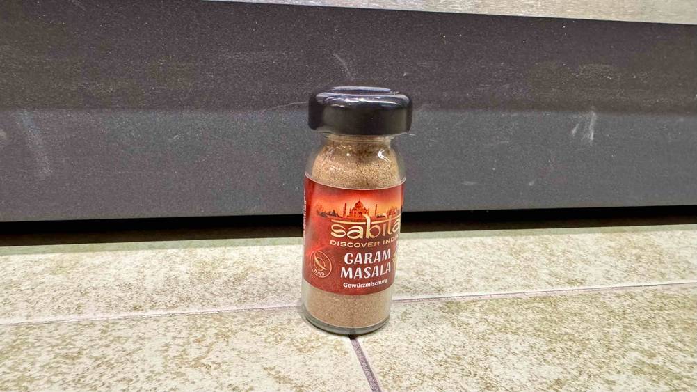Vor einer grauen Leiste steht ein Glas Garam Masala im Test auf dem Boden.