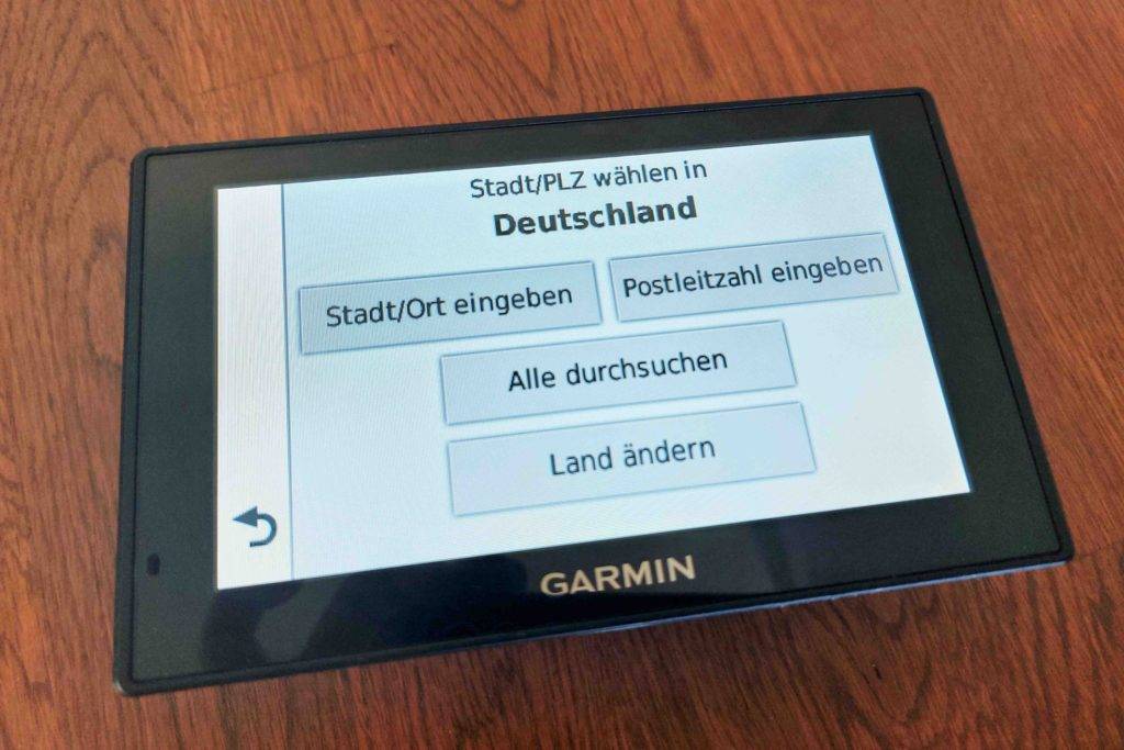 Im Test: Garmin Navi liegt mit angezeigtem Eingabedisplay für Stadtauswahl auf einem Holzboden.