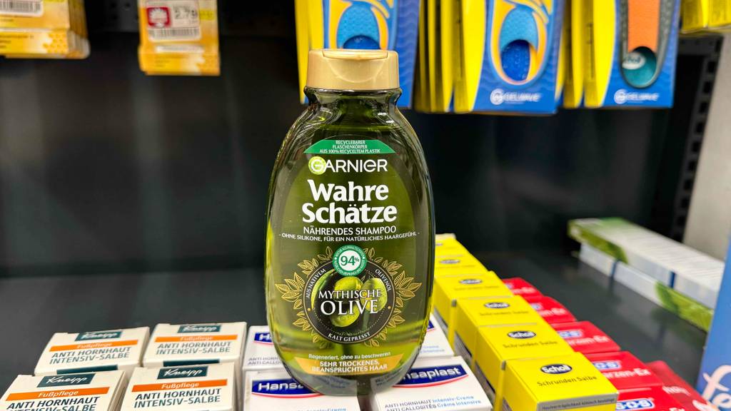 Eine grün-transparente Kunststoffflasche mit goldenem Spenderverschluss inklusive Olivenöl-Shampoo im Test steht auf Hautpflegemitteln im Drogeriemarkt.