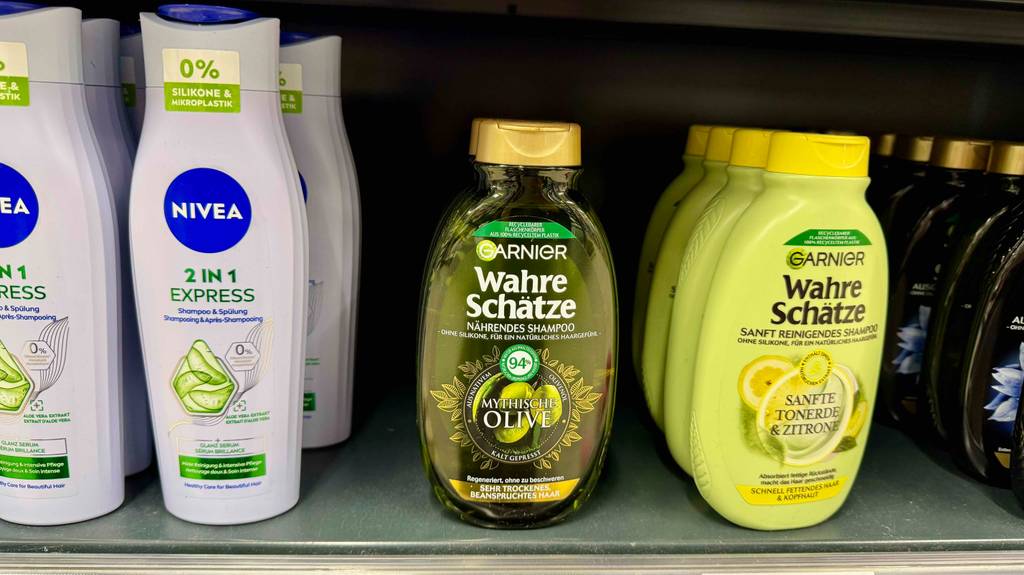 Eine grün-transparente Quetschflasche mit Olivenöl-Shampoo im Test steht neben anderen Shampoosorten in einem Verkaufsregal.