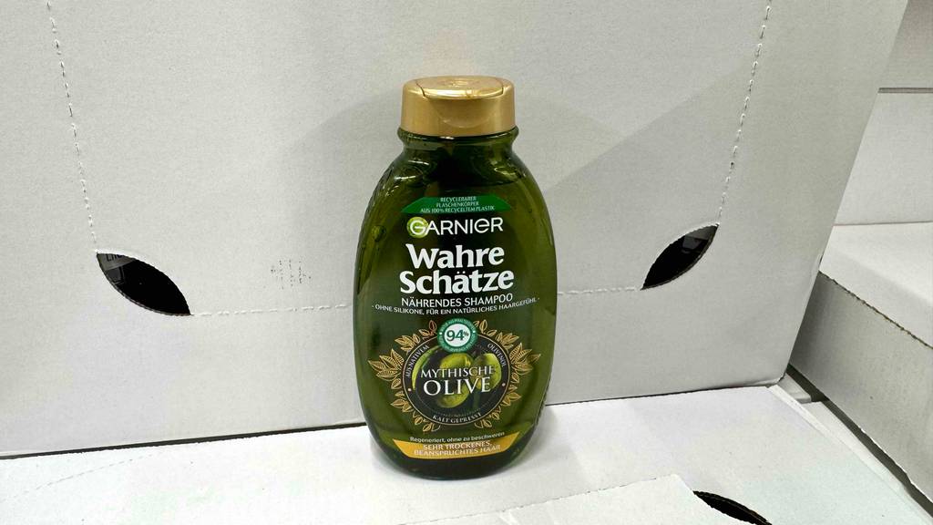Das getestete Olivenöl-Shampoo befindet sich in einer grün-transparenten Kunststoffflasche mit goldenem Spenderverschluss auf einem weißen Karton.