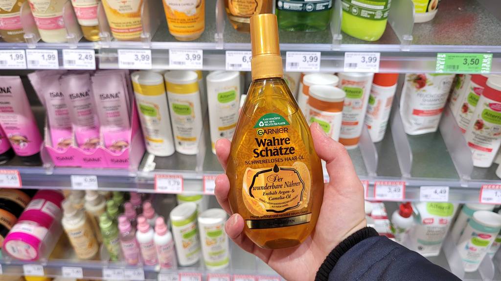 Hand hält eine goldfarbene Garnier Flasche mit Haaröl im Test vor einem Verkaufsregal mit vielen verschiedenen Pflegeprodukten.