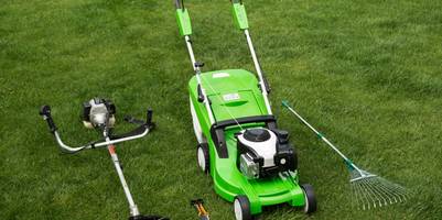 Ein Rasenmäher. ein Rasentrimmer und ein Rechen nebeneinander auf dem Rasen liegend.