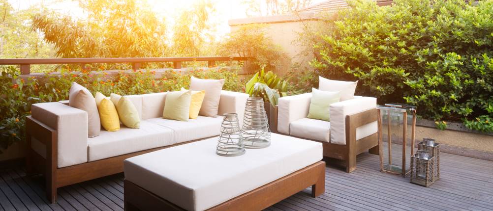Terrasse mit Gartenmöbeln und hellen Polstern, umgeben von Sträuchern und Bäumen.