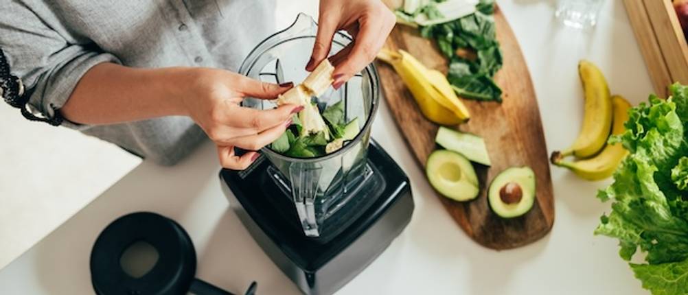 Standmixer getestet: Ein Standmixer ist von oben zu sehen. Zwei Frauenhände füllen Bananen in den Mixbehälter, in dem sich bereits weitere Zutaten befinden. Bananenschalen, Avocados und weiteres Gemüse liegt auf einem Holzbrett neben dem Mixer.