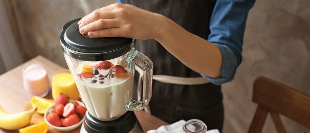 Gastroback-Mixer-Test: Eine Hand hält den schwarzen Deckel eines Glas-Standmixers fest. In dem Mixer befinden sich Milch und Obst. Weitere Zutaten liegen auf der Arbeitsfläche um den MIxer herum.