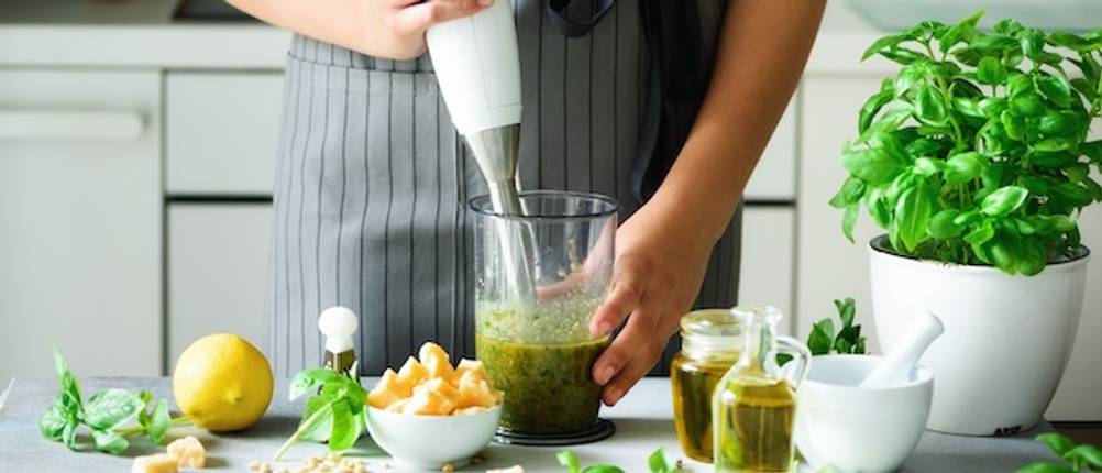Gastroback-Mixer gestestet: Eine Frau bedient einen weißen Stabmixer und mix in einem hohen Gefäß grünes Gemüse für ein Pesto. Weitere Zutaten stehen auf der Ablagefläche.