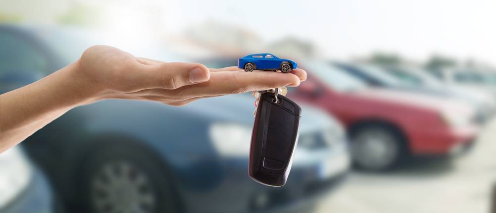 Gebrauchtwagen-kaufen-Test: Eine Person hält ein blaues Modellauto und einen Autoschlüssel in der Hand, während im Hintergrund einige Autos parken.