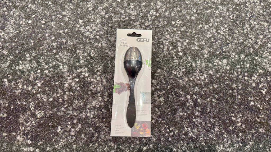 Getestet: Ein Tee-Ei im Löffeldesign liegt in der Verpackung auf einem grau melierten Teppich.