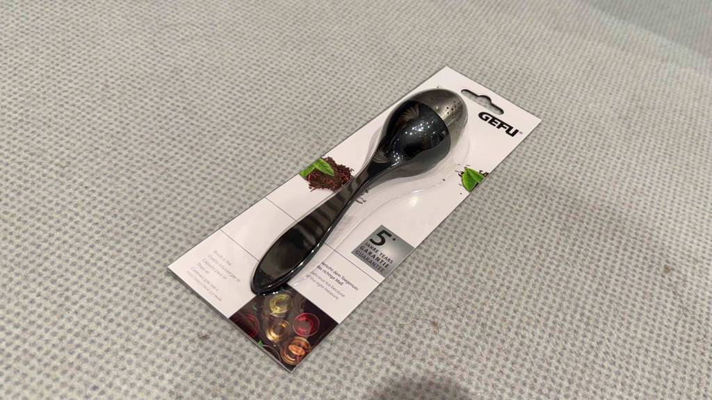 Ein getestetes Tee-Ei im Löffeldesign liegt in seiner Verpackung auf einem hellen Untergrund.