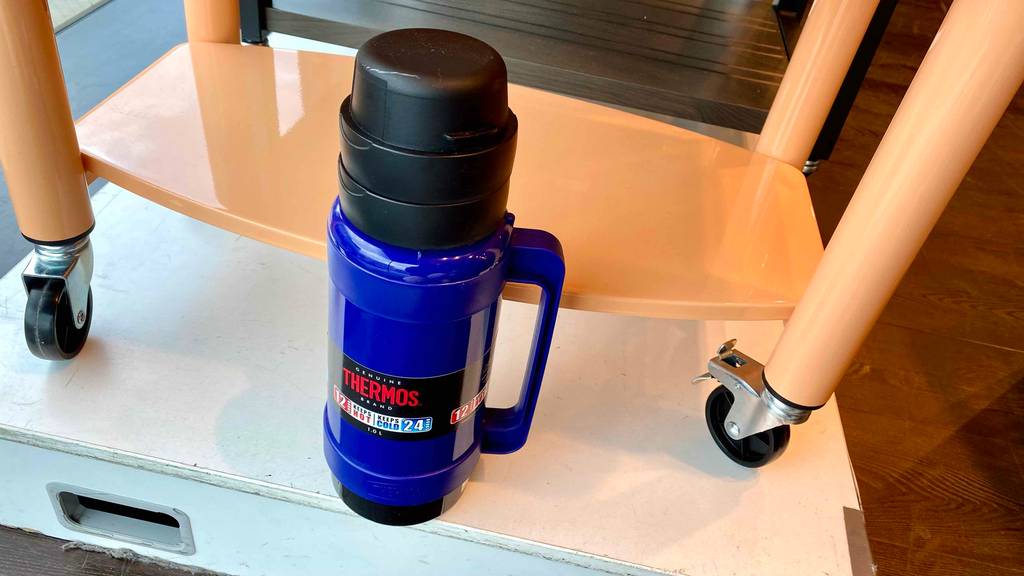 Eine getestete Outdoor-Thermoskanne in Blau mit Handgriff, schwarzem Verschluss und Trinkbecher steht vor einem weißen Serviertisch inklusive Rollen.