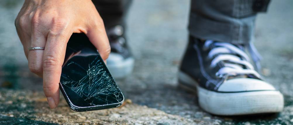 Geräteversicherungen im Test: Eine Person in Stoffschuhen hebt ein Smartphone mit zerbrochenem Display auf.