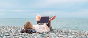 Geräteversicherungen im Test: Eine junge Frau liegt an einem Kiesstrand, während sie ihren Laptop auf den Beinen hat.