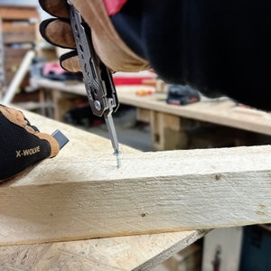 Zwei Hände mit Arbeitshandschuhen nutzen das Gerber Multitool, um eine Schraube in ein Holzbrett zu drehen. Der Hintergrund zeigt eine Werkstatt mit Werkzeugen.