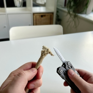 Zwei Hände halten ein Gerber Multitool Truss mit ausgeklappter Klinge und ein ausgefranstes Seil. Der Hintergrund zeigt einen weißen Tisch mit einer hellen, modernen Einrichtung.