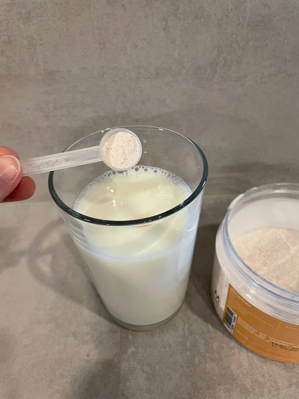 Eine Hand hält einen Löffel des getesteten Geschmackspulvers über ein Glas mit Milch neben der Dose mit Pulver.