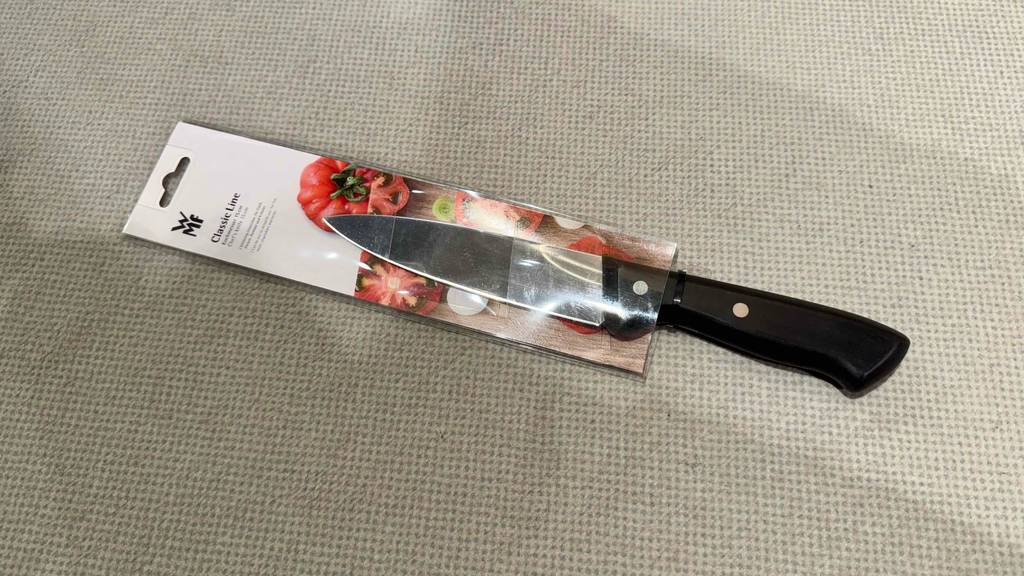 Ein getestetes WMF-Kochmesser mit schwarzem Griff liegt innerhalb der Verpackung auf einer hellgrauen Oberfläche.
