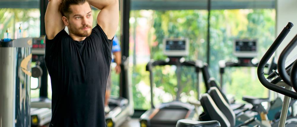 Gewichtheberschuhe für Herren im Test: Ein junger Mann wärmt sich vor dem Einsatz an den Geräten in einem Fitnessstudio auf, indem er Stretching-Übungen durchführt.