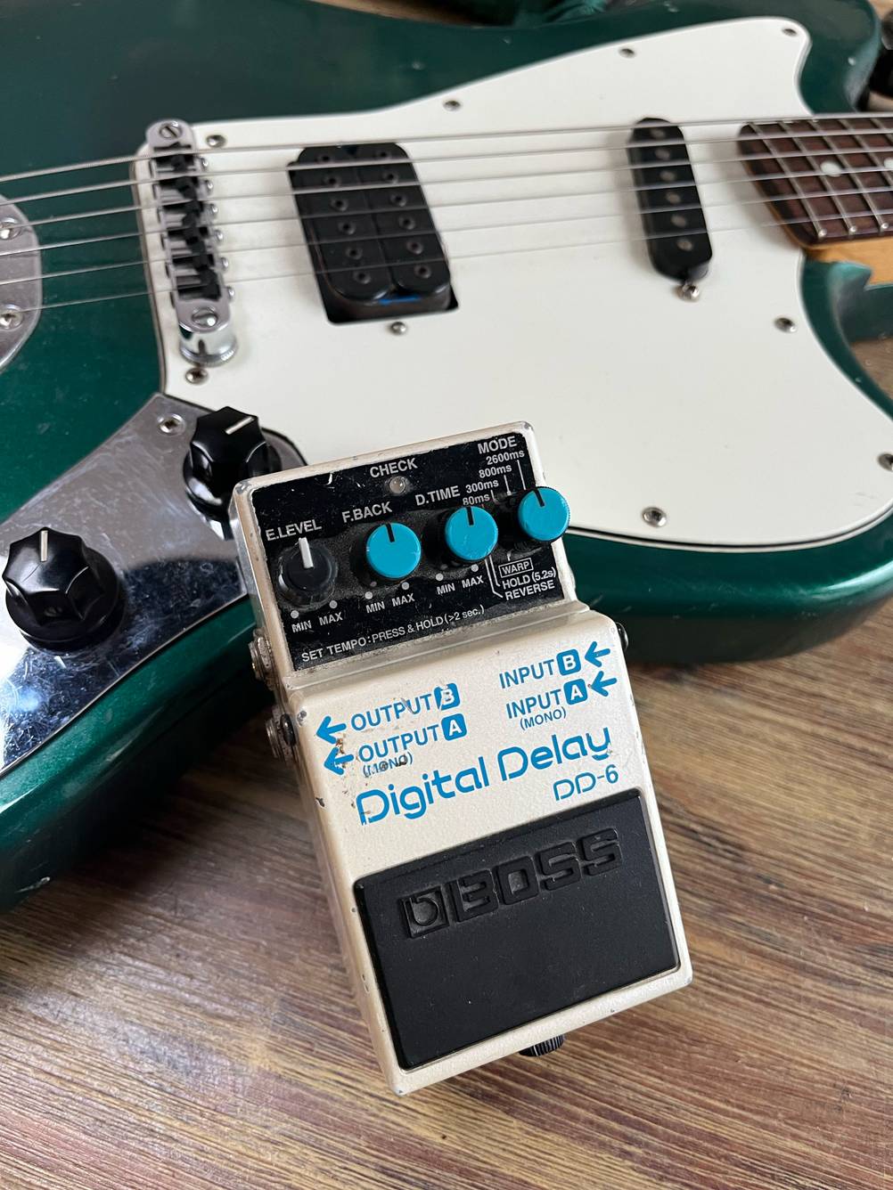 Ein getestetes Gitarren-Effektgerät steht angelehnt an einer Gittare auf dem Boden.