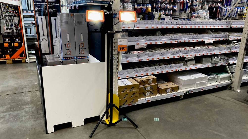Im Test: Ein LED-Baustrahler mit Stativ steht in einem Baummarkt vor einem Regal mit Produkten.