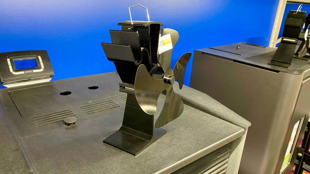 Ein Ofenventilator im Test auf einem Ofen stehend schräg von der Seite vor einer blauen Wand.