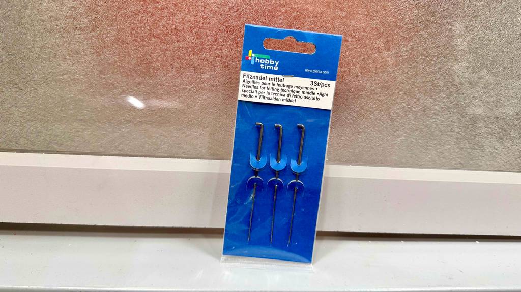 Filznadeln getestet: Eine blaue Packpappe mit drei Filznadeln steht vor einer beigfarbenen Wand.