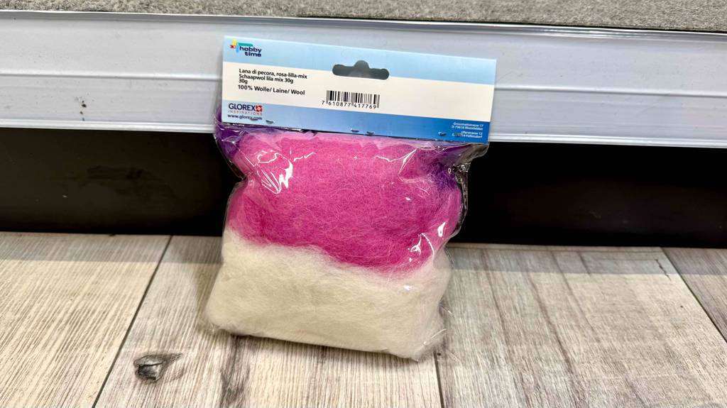 Die Filzwolle im Test ist weiß, rosa und lila und befindet sich in der 30-g-Packung vor einem weißen Regal.